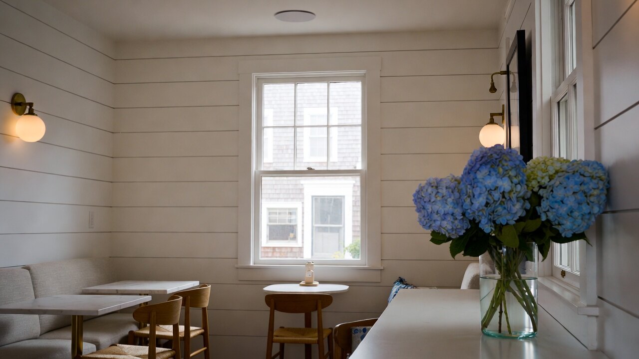 Calm, tastefully appointed coastal café interior with shiplap walls, marble tables, brass sconces, blue hydrangeas, and a discreet architectural in-ceiling speaker — the kind of tasteful background-music setup AES Integration installs for small businesses