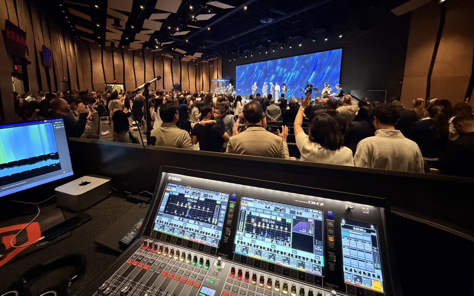 Front-of-house view at an AES Integration church upgrade in Massachusetts — a Yamaha DM7 digital console with a Mac mini running real-time room measurement software during a live worship service, congregation standing with hands raised in front of a full LED video wall